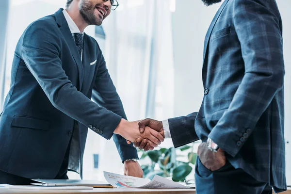 Обрезанный образ улыбающихся мультикультурных бизнесменов, пожимающих руки в офисе — стоковое фото