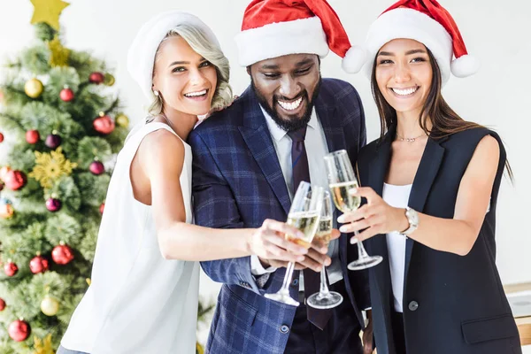 Empresários multiétnicos felizes em chapéus santa clinking com copos de champanhe no escritório — Fotografia de Stock