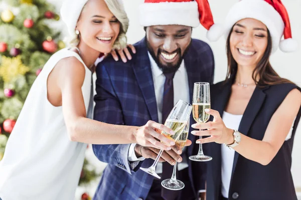 Sorridente multiétnico empresários em santa chapéus clinking com copos de champanhe no escritório — Fotografia de Stock