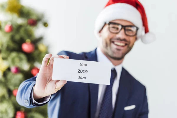 Homem de negócios sorridente em cartão de retenção de santa hat com 2019 no escritório — Fotografia de Stock