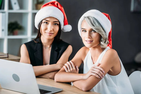 Attraktive multikulturelle Geschäftsfrauen mit Weihnachtsmannhüten, die im Büro in die Kamera schauen — Stockfoto