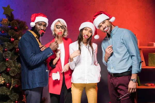 Multiethnic managers holding lips and glasses on sticks at new year corporate party — Stock Photo