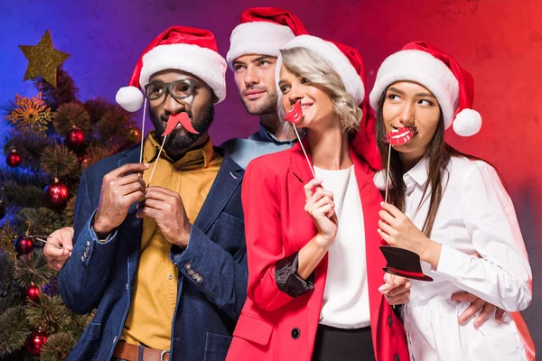 Empresarios multiculturales sosteniendo los labios y gafas en palos en la fiesta corporativa de año nuevo y mirando hacia otro lado - foto de stock