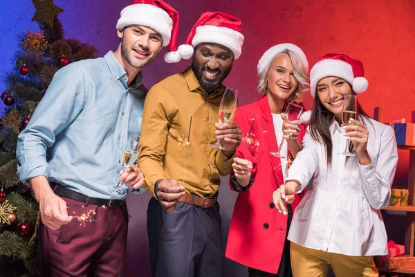 Young multicultural businesspeople holding christmas sparklers and glasses of champagne at new year corporate party — Stock Photo