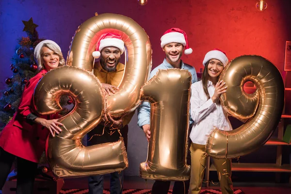 Jovens empresários multiétnicos em santa chapéus segurando 2019 balões na festa corporativa do ano novo — Fotografia de Stock