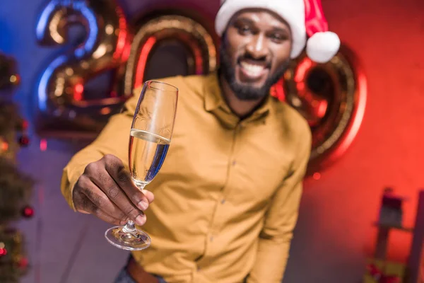 Happy african american businessman in santa hat showing glass of champagne at new year corporate party — Stock Photo