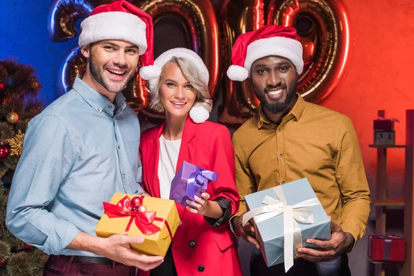 Heureux entrepreneurs multiculturels dans santa chapeaux tenant des boîtes-cadeaux à la nouvelle fête d'entreprise de l'année — Photo de stock