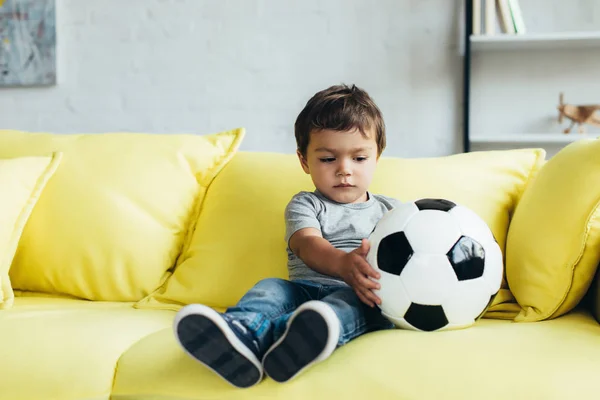 Menino bonito sentado no sofá amarelo com bola de futebol — Fotografia de Stock