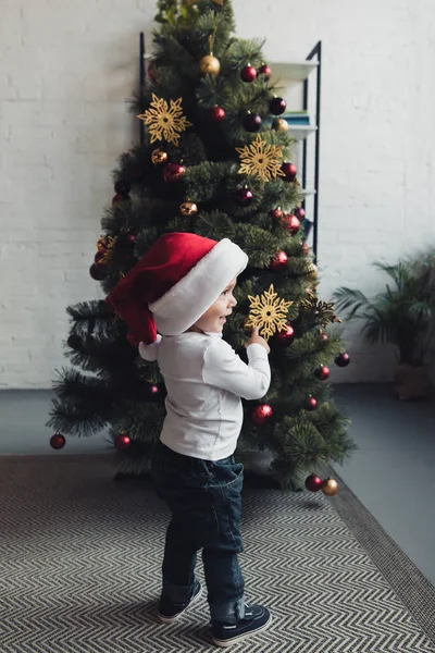 Süßer lächelnder Junge mit Weihnachtsmütze, der auf den Weihnachtsbaum zeigt — Stockfoto