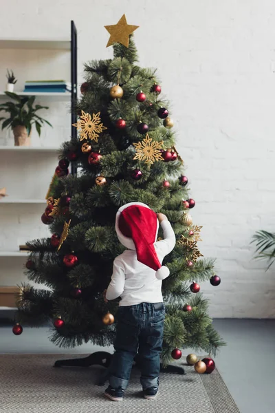 Vue arrière de l'enfant en chapeau de Père Noël décorant l'arbre de Noël à la maison — Photo de stock