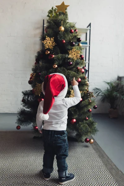 Visão traseira da criança masculina em santa hat apontando para a árvore de natal — Fotografia de Stock