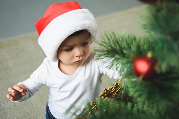 Niedlicher Junge mit rotem Weihnachtsmann-Hut schmückt den Weihnachtsbaum — Stockfoto