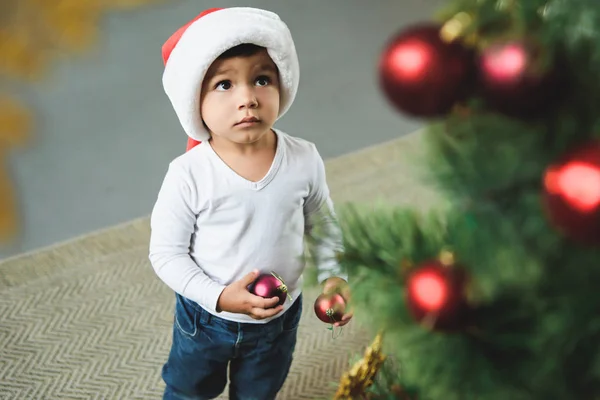 Adorable child in santa hat decorating christmas tree with balls — Stock Photo