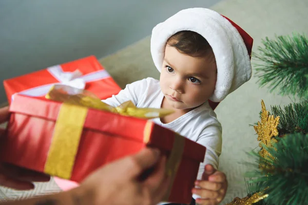 Cute son in santa hat taking red gift box from parents hands near christmas tree — Stock Photo