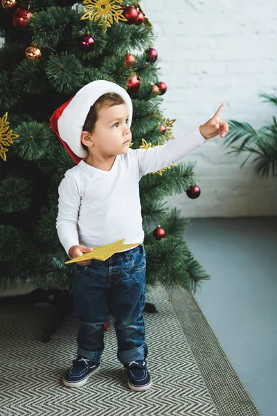 Adorable boy in santa hat holding golden star while pointing up and standing near christmas tree — Stock Photo