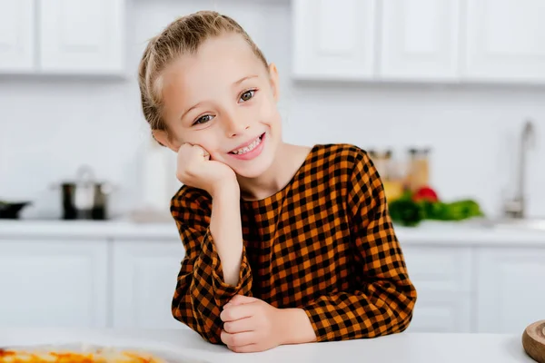 Adorável criança se inclinando na mesa na cozinha e olhando para a câmera — Fotografia de Stock