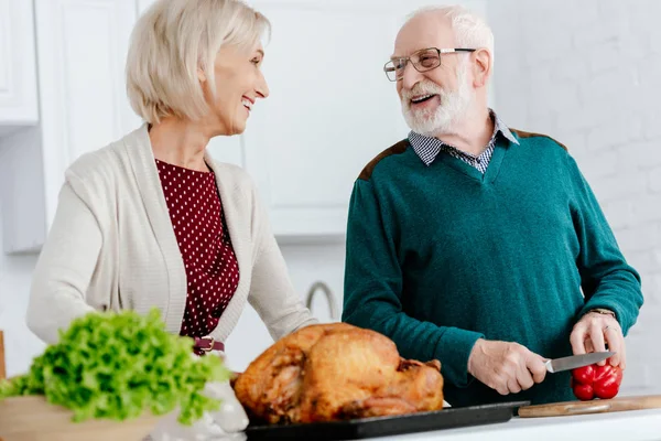 Laughing senior couple cooking thanksgiving turkey together and looking at each other — Stock Photo