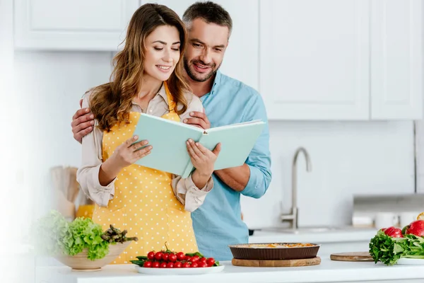 Счастливая пара чтение рецепт книги во время приготовления пищи вместе на кухне — стоковое фото