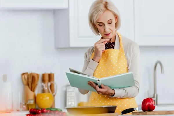 Mujer mayor leyendo libro de recetas mientras cocina en la cocina - foto de stock