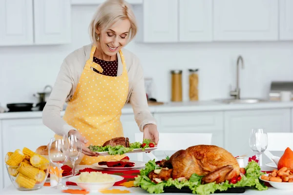 Smiling senior woman carrying meal on table with delicious dishes for thanksgiving celebration — Stock Photo