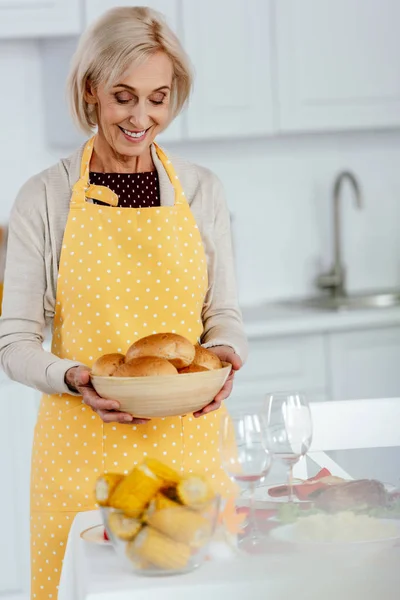 Feliz mulher sênior segurando tigela com pão perto da mesa servida para o jantar de ação de graças — Fotografia de Stock