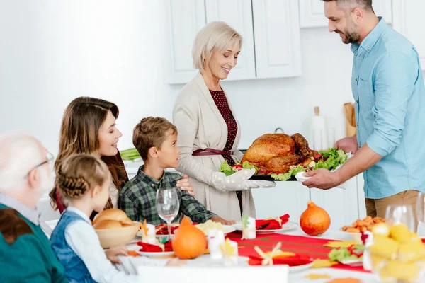 Femme âgée et homme adulte portant la dinde cuite au four pour le dîner de Thanksgiving avec la famille à la maison — Photo de stock