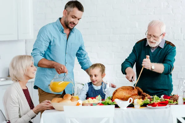Großvater schneidet Truthahn, während erwachsener Mann Saft an serviertem Tisch für Erntedankfeier mit Familie einschenkt — Stockfoto