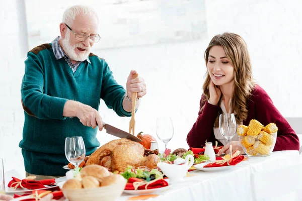 Heureux senior homme tranchant délicieuse dinde pour Thanksgiving célébration tandis que sa fille adulte assis près de la table — Photo de stock