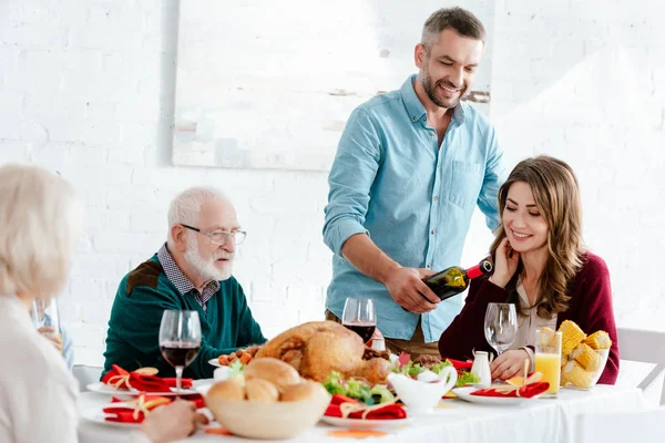 Счастливый взрослый мужчина наливает вино в бокал за сервированным столом, в то время как семья празднует День Благодарения — стоковое фото