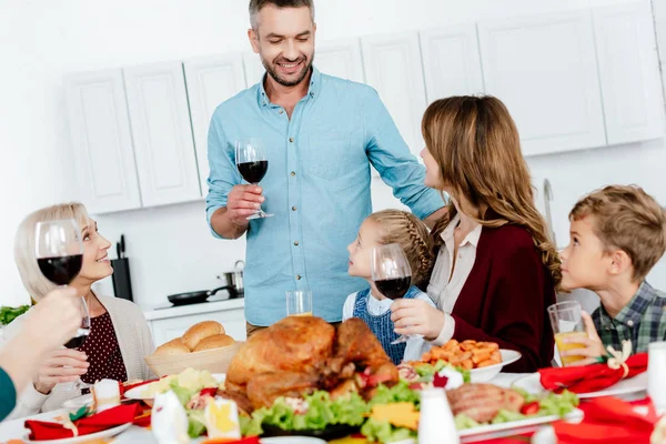 Heureux homme adulte avec verre de vin faire du pain grillé à la table servie avec de la dinde tandis que sa famille célèbre thanksgiving — Photo de stock