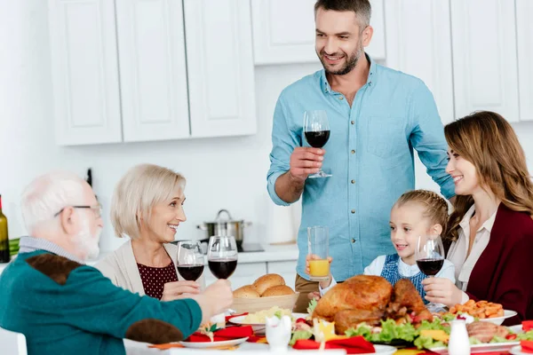 Hombre adulto con copa de vino haciendo tostadas mientras su familia celebra la acción de gracias en la mesa servida - foto de stock