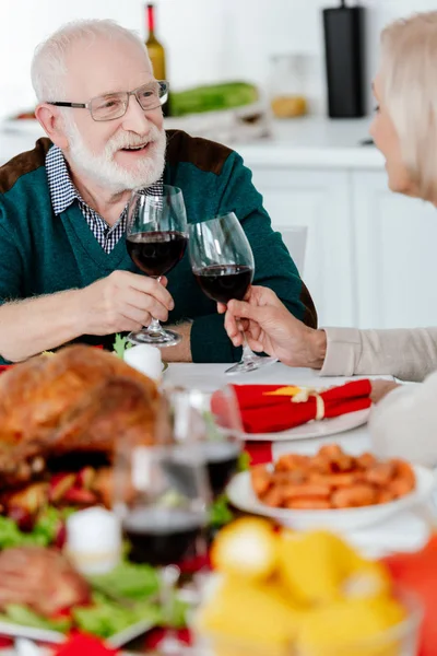 Sonriente pareja mayor tintineo por copas de vino en servir mesa con pavo al horno en acción de gracias - foto de stock