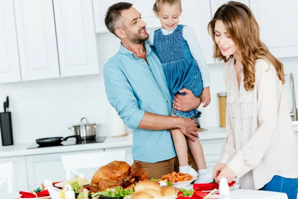 Adulto feliz homem segurando filha enquanto sua esposa servindo mesa para jantar de ação de graças — Fotografia de Stock