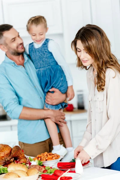 Sonriente hombre adulto sosteniendo hija mientras su esposa sirviendo mesa para la celebración de acción de gracias - foto de stock