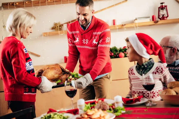 Smiling man and woman carrying turkey for holiday dinner while little kid in christmas hat sitting on grandfather knees at table — Stock Photo