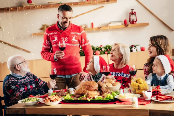 Hombre adulto feliz sosteniendo copa de vino y haciendo tostadas durante la celebración de Navidad con la familia en casa - foto de stock