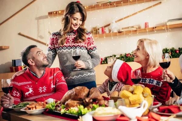 Hermosa mujer sonriente con copa de vino haciendo tostadas durante la cena de Navidad con la familia feliz en casa - foto de stock
