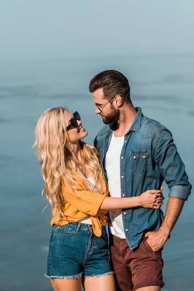 Couple in sunglasses hugging and looking at each other near sea — Stock Photo