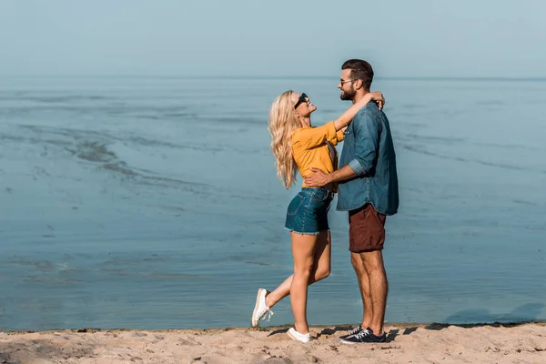 Pareja en gafas de sol abrazándose y mirándose en la playa - foto de stock