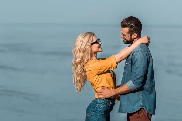 Pareja en gafas de sol abrazándose y mirándose el uno al otro cerca del océano - foto de stock