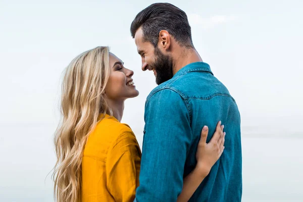 Vista lateral de la pareja sonriente abrazándose y mirándose el uno al otro contra el cielo azul - foto de stock