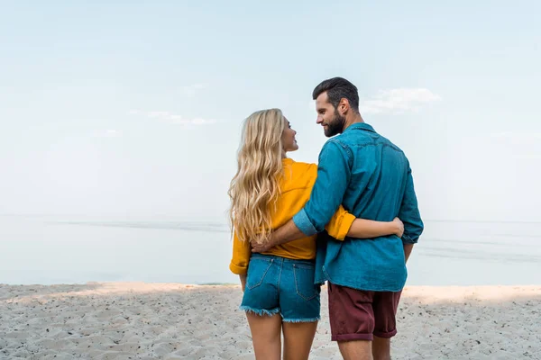 Вид ззаду пара обіймається, ходить і дивиться один на одного на пляжі — стокове фото