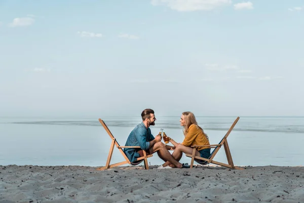 Vista laterale di coppia seduta su lettini e clinking con bottiglie di birra di vetro sulla spiaggia sabbiosa — Foto stock