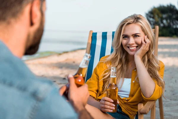 Pareja sentada en tumbonas con cerveza en botellas en la playa de arena, novia sonriente mirando novio - foto de stock