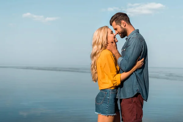 Vista lateral de novia sonriente y novio tocando con narices en la playa - foto de stock