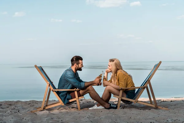 Vista lateral de la novia y el novio sentado en tumbonas y tintineo con botellas de vidrio de cerveza en la playa de arena - foto de stock