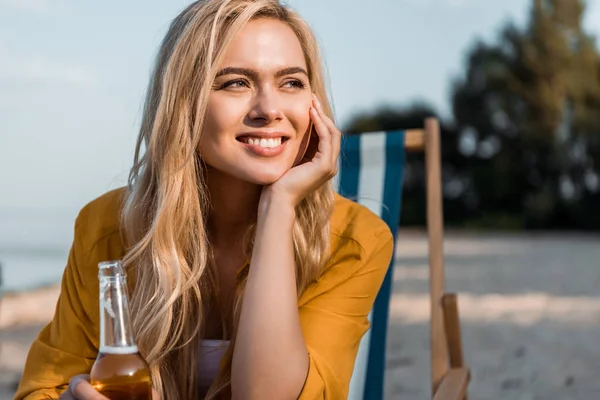 Attraktives lächelndes Mädchen, das mit Bier in der Flasche auf einer Sonnenliege sitzt und wegschaut — Stockfoto