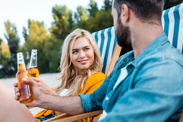 Couple sitting on sun loungers and clinking with glass bottles of beer on sandy beach — Stock Photo