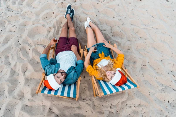 Vista de alto ângulo de casal em chapéus de santa deitado em espreguiçadeiras com cerveja em garrafas na praia arenosa — Fotografia de Stock