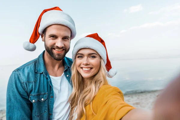 Punto de vista de la cámara sonriente pareja en sombreros de santa mirando a la cámara en la playa - foto de stock